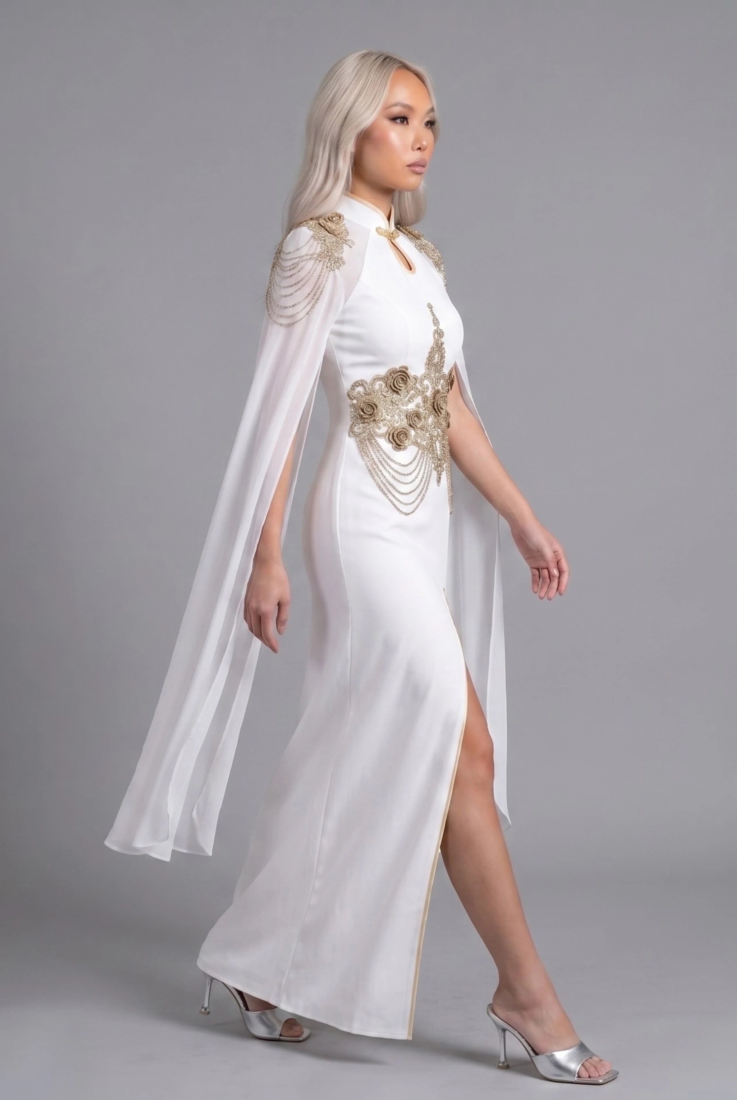 Side profile view of a woman walking in the white fusion dress, showcasing the high side slit and dramatic movement of the cape