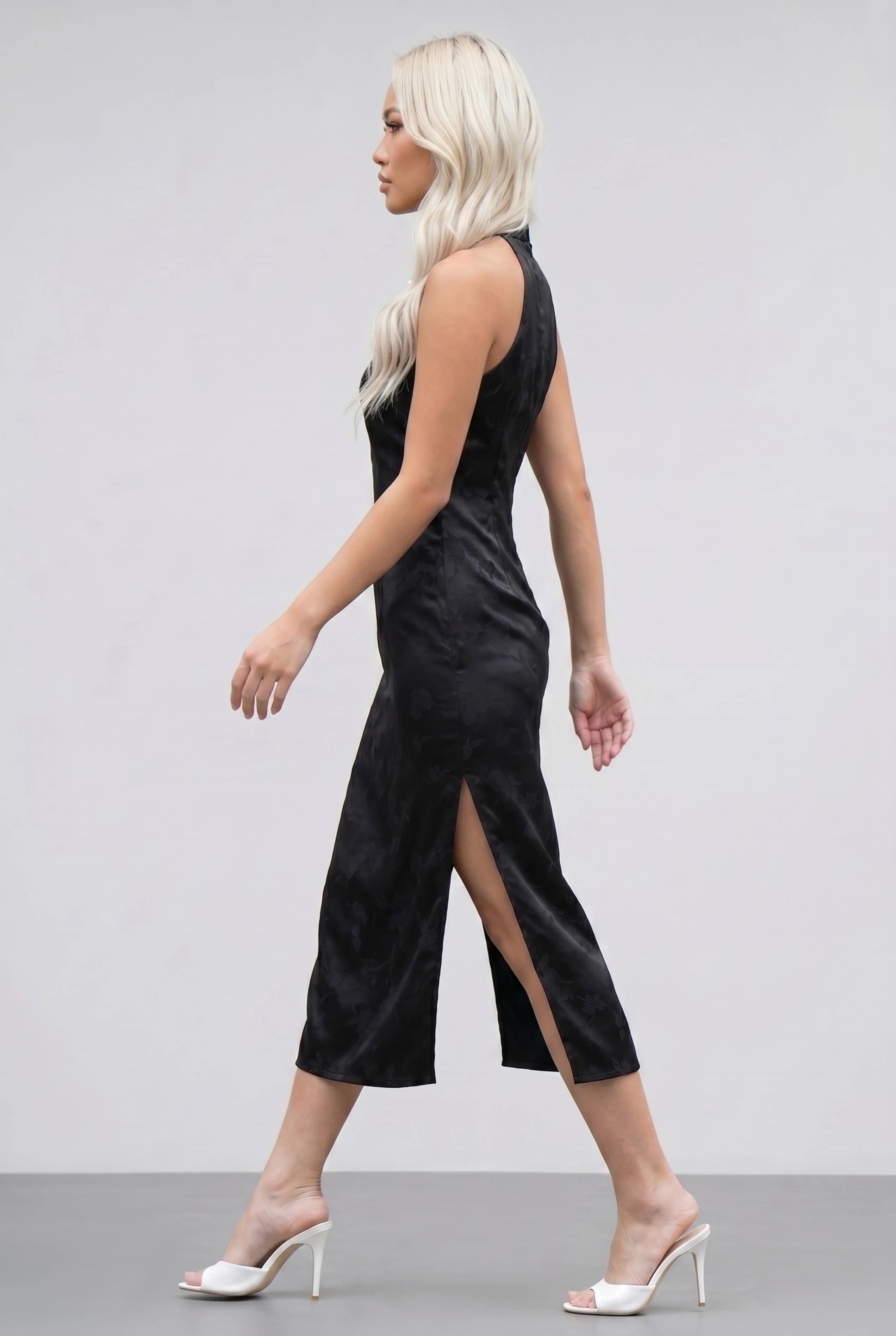 Full-body side profile view of a woman in stride wearing the black qipao with a tonal floral pattern and white heels