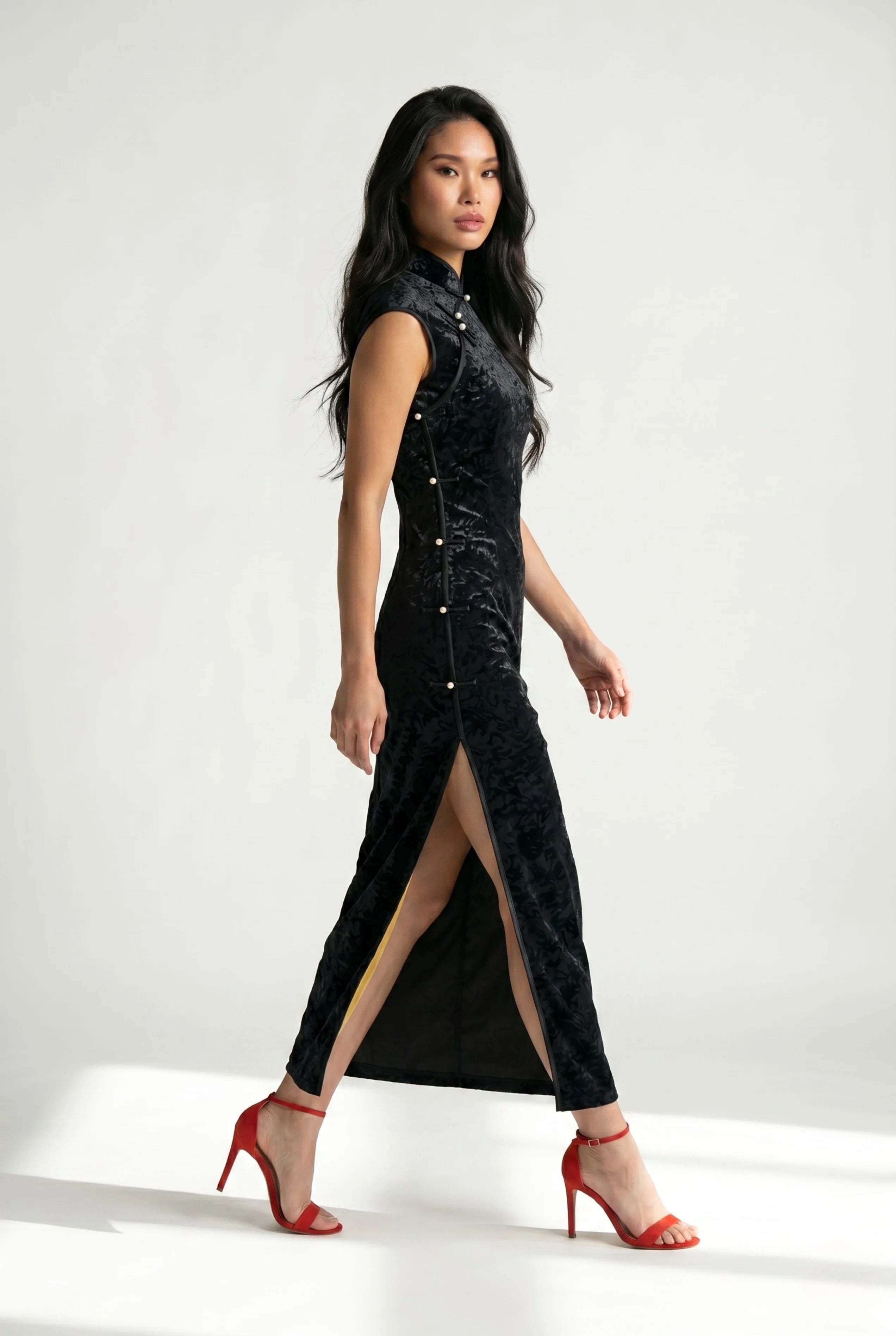 Side profile view of a woman walking in a black velvet cheongsam, showcasing the high slit and red heels.