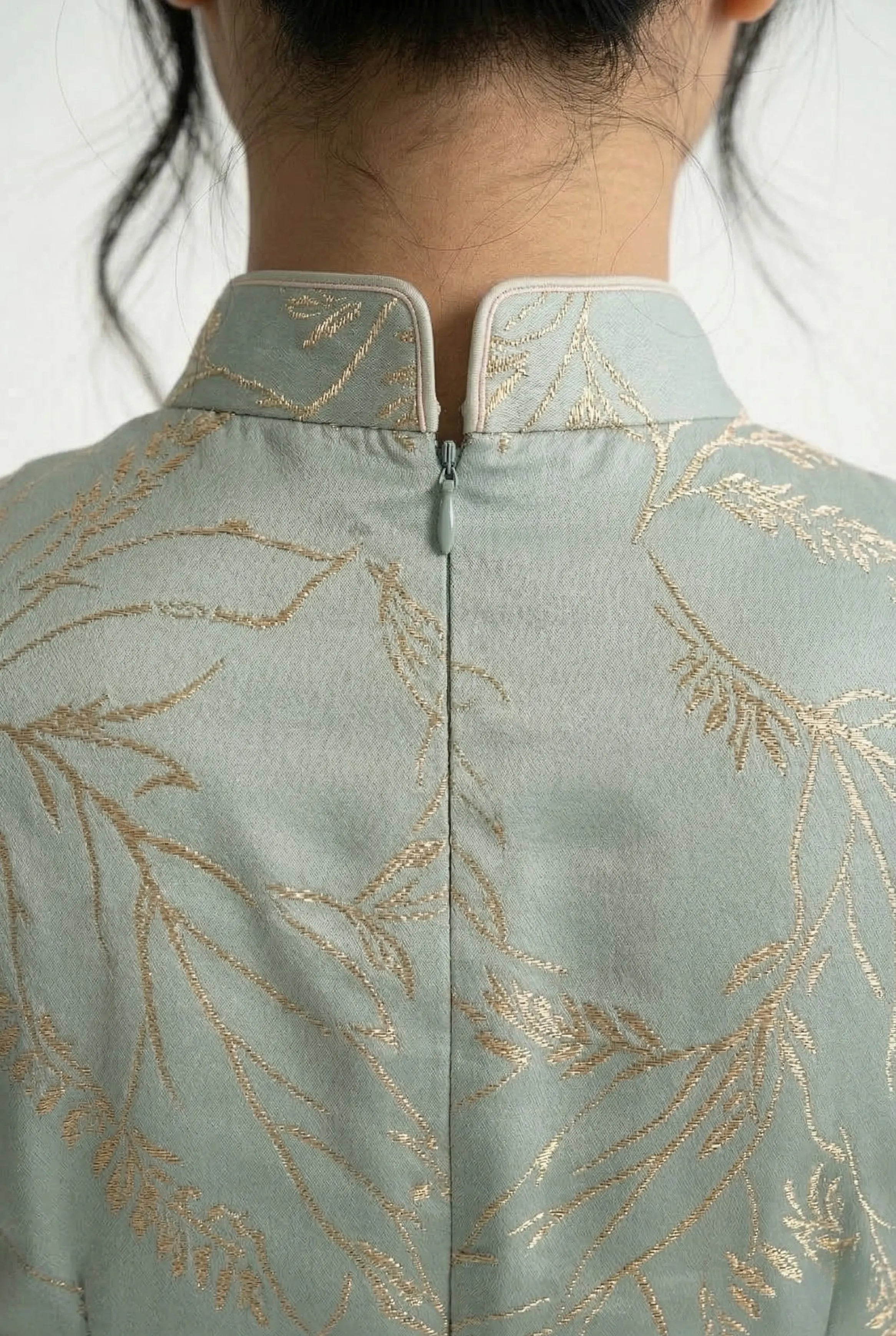 Detailed shot of the high back mandarin collar and neckline of a sage blue Qipao, showcasing the gold embroidery.