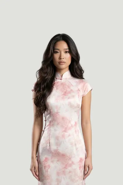 A model wearing a blush pink silk qipao by Maison Hua, featuring a soft watercolor floral print, cap sleeves, and a traditional mandarin collar for a romantic, modern look