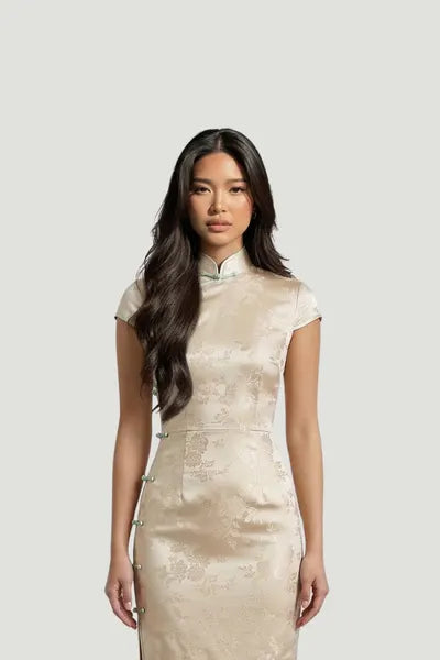 A model showcasing a champagne-colored silk qipao by Maison Hua, featuring a delicate floral brocade pattern, cap sleeves, and a classic mandarin collar