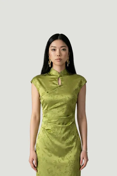 A model wearing an olive green silk qipao by Maison Hua, featuring a subtle floral pattern, elegant cap sleeves, and a traditional Mandarin collar