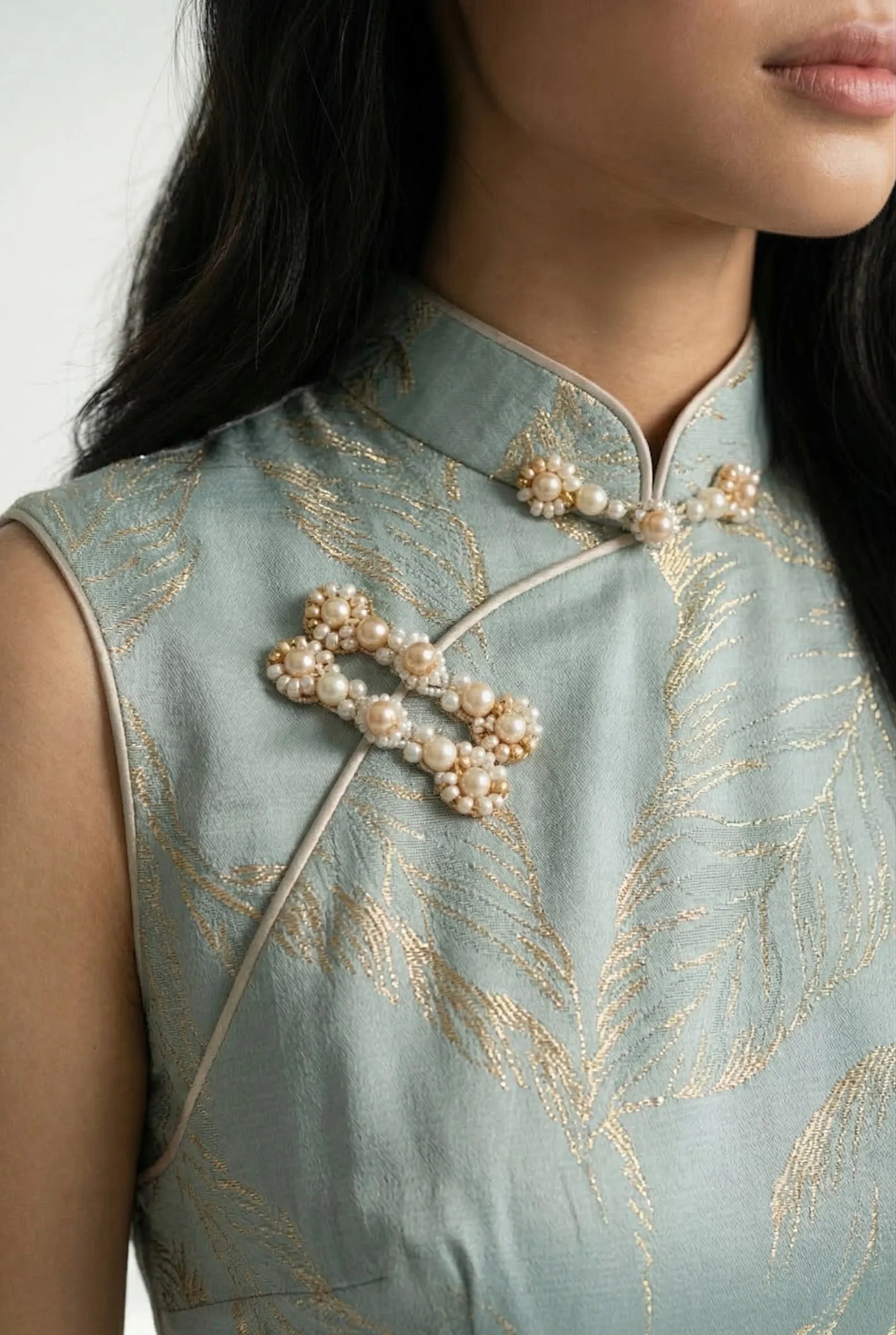 Close-up of the traditional mandarin collar and beaded frog button closure on a sage blue Qipao with gold leaf motifs.