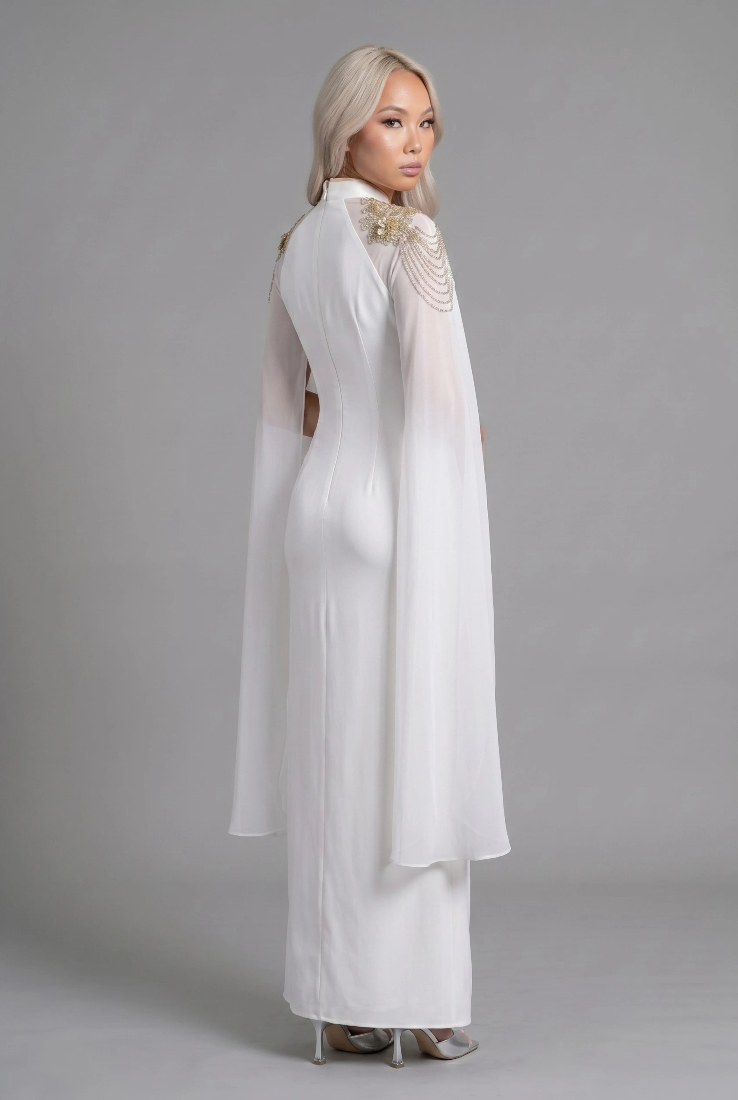 Three-quarter back profile view highlighting the flowing sheer cape sleeves and form-fitting cut of the white and gold dress